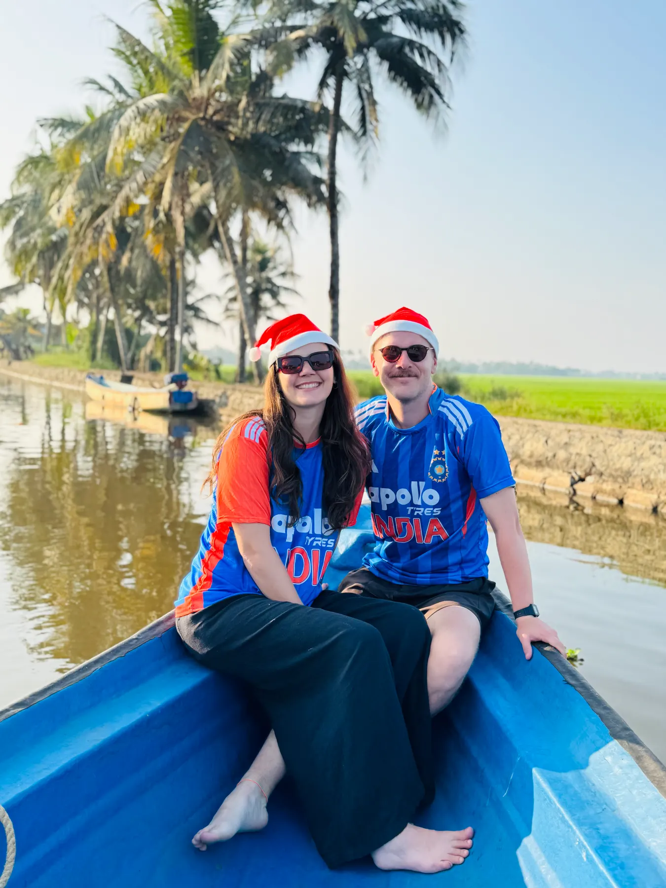 Two guests in Santa hats smiling in the bow of a blue boat beside green paddy fields and palms