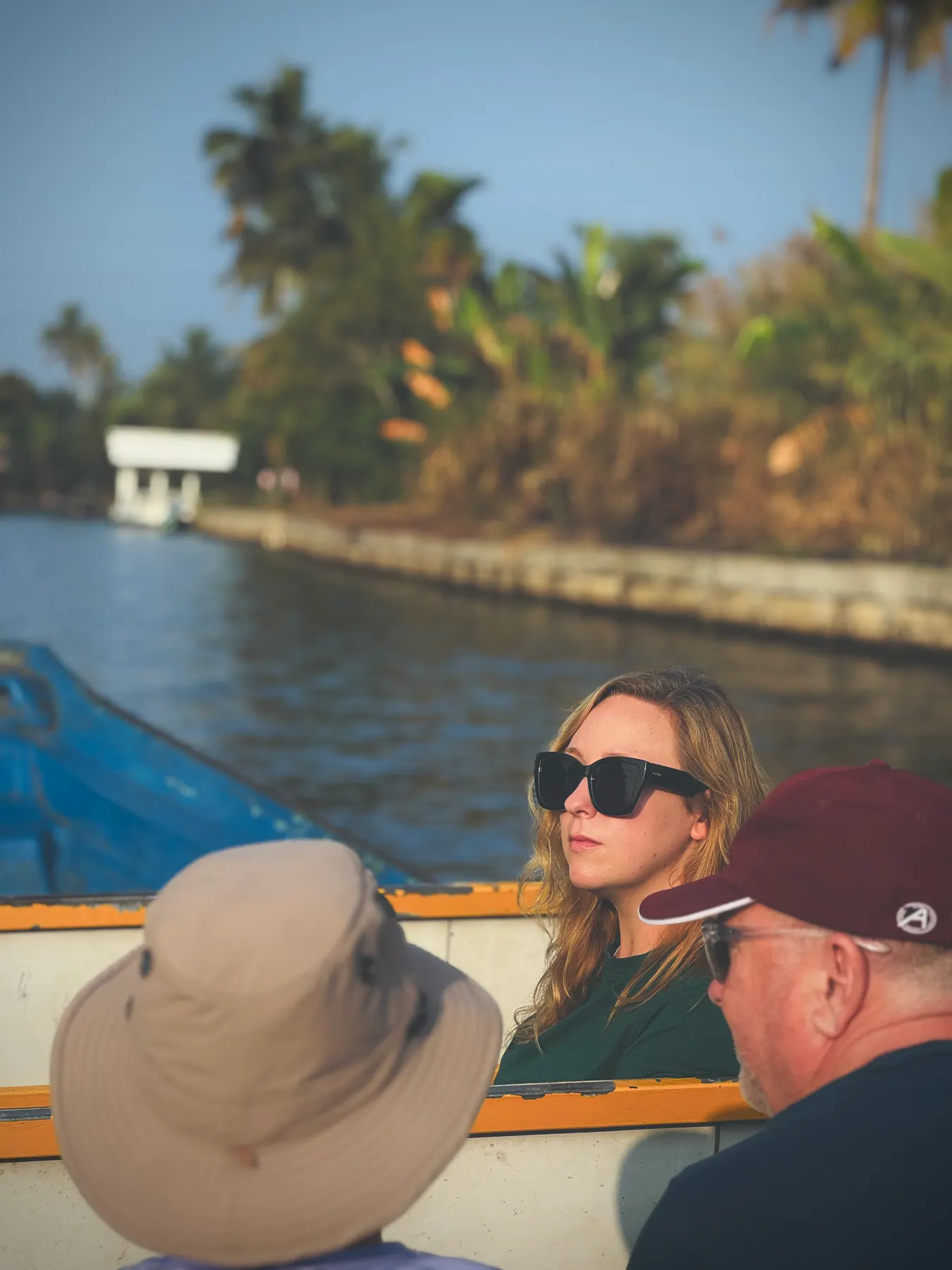 A guest in sunglasses seated in a boat in warm evening light, with a canal bank and palms behind