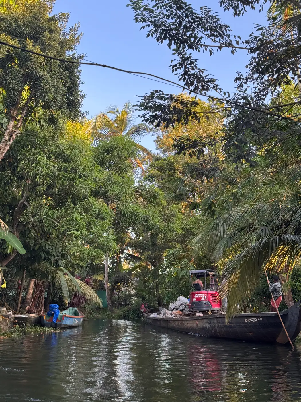 A canal bordered by dense green trees, with a loaded village boat ahead and its reflection in the water