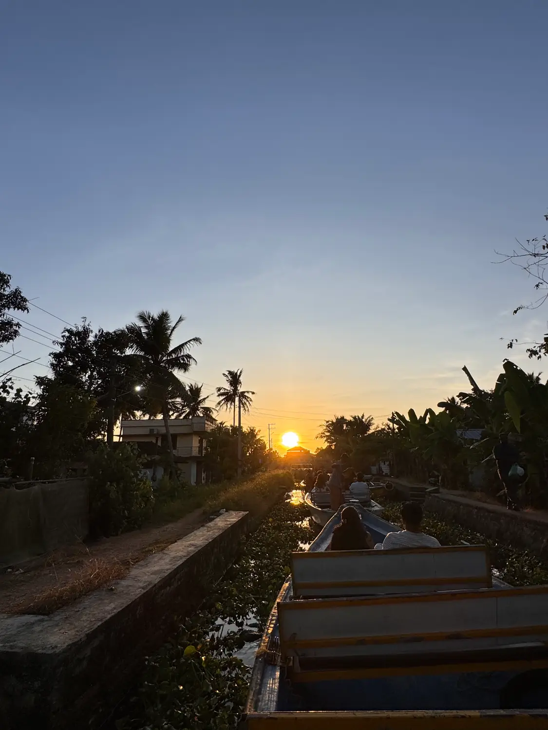 A canal leading toward a low orange sun beneath a clear sky, with silhouetted palms and passengers in the bow of a boat