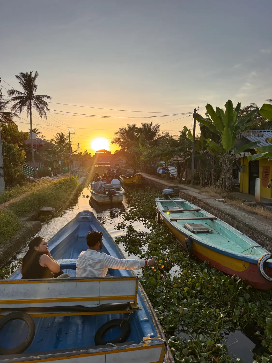 Passengers in a boat on a narrow canal toward the setting sun, with a colourful moored boat and palms on the bank