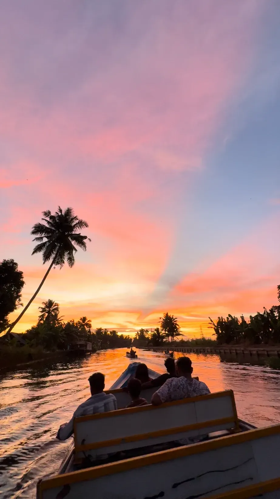 Passengers silhouetted in a boat heading down a canal toward a pink and orange sunset, with a leaning coconut palm