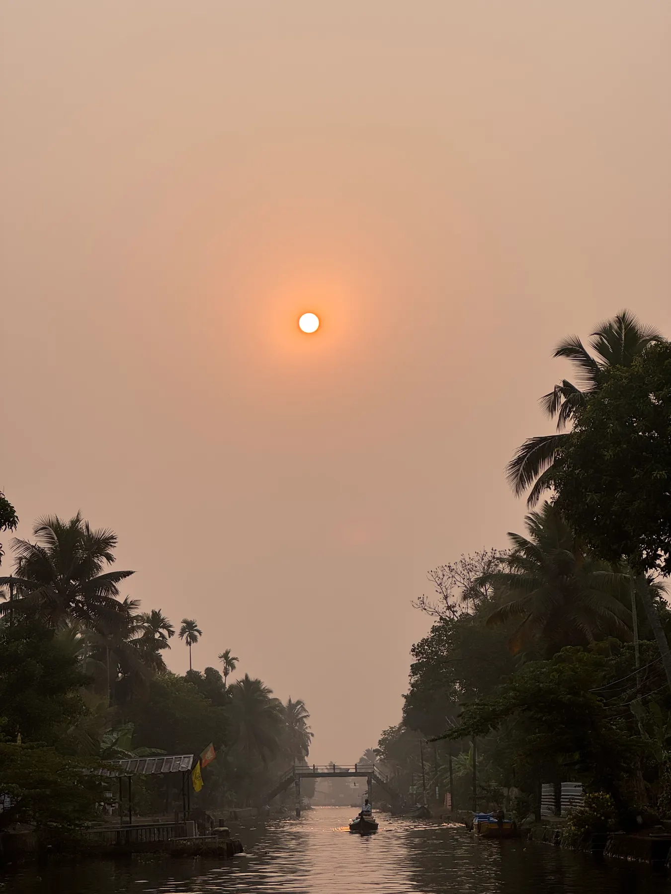 A small boat on a canal beneath a pale sun in a hazy orange sky, with a footbridge and palms beyond