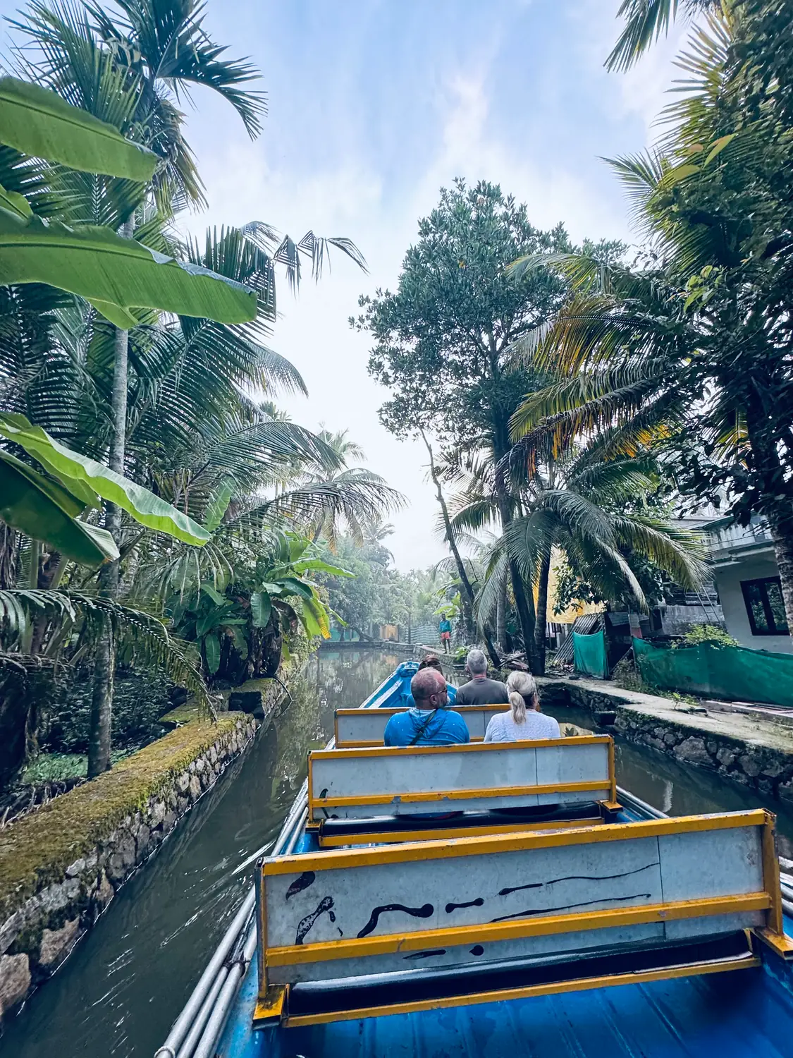 Passengers gliding down a narrow canal lined with palms, banana leaves and a moss-covered stone wall