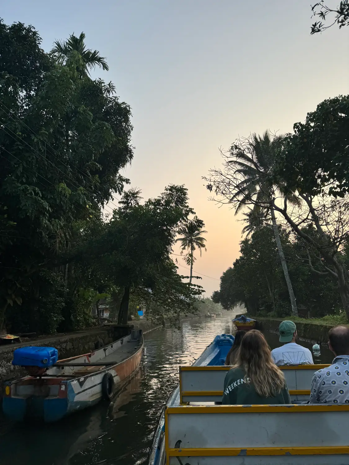 A boat with passengers heading up a tree-lined canal toward a soft peach evening sky