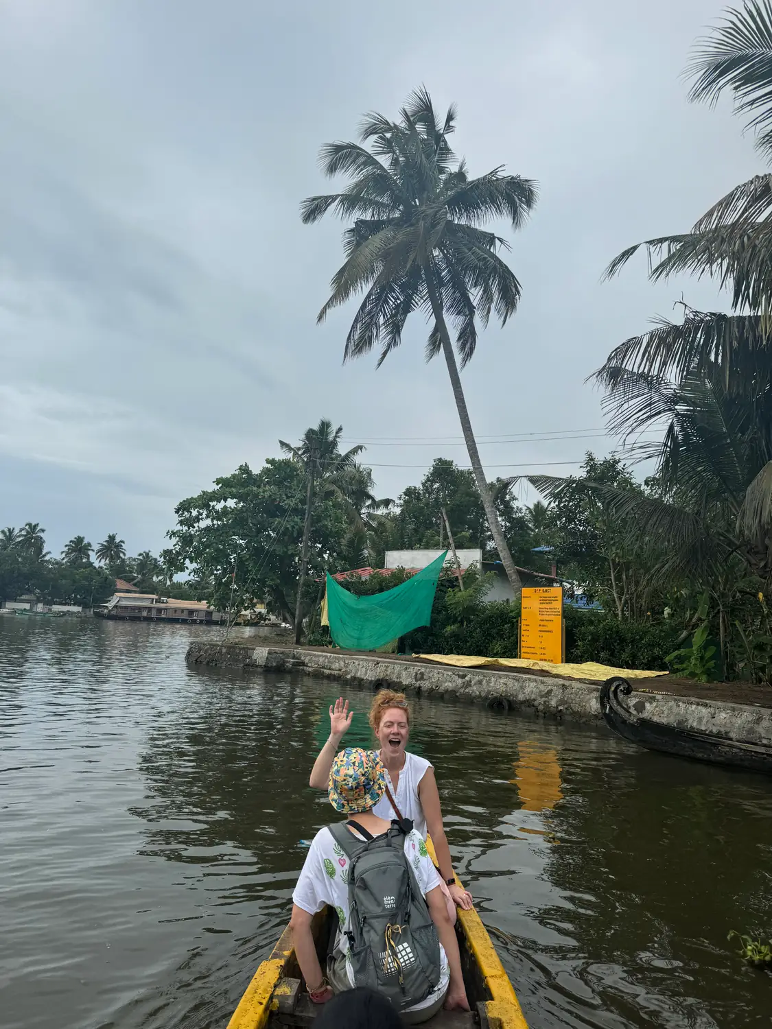 A guest waving and laughing from a boat on a backwater canal, with a leaning coconut palm behind