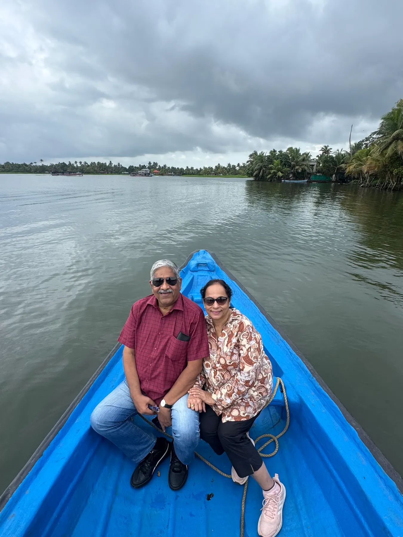 Two guests seated in the bow of a blue boat on a wide, calm backwater beneath heavy monsoon clouds