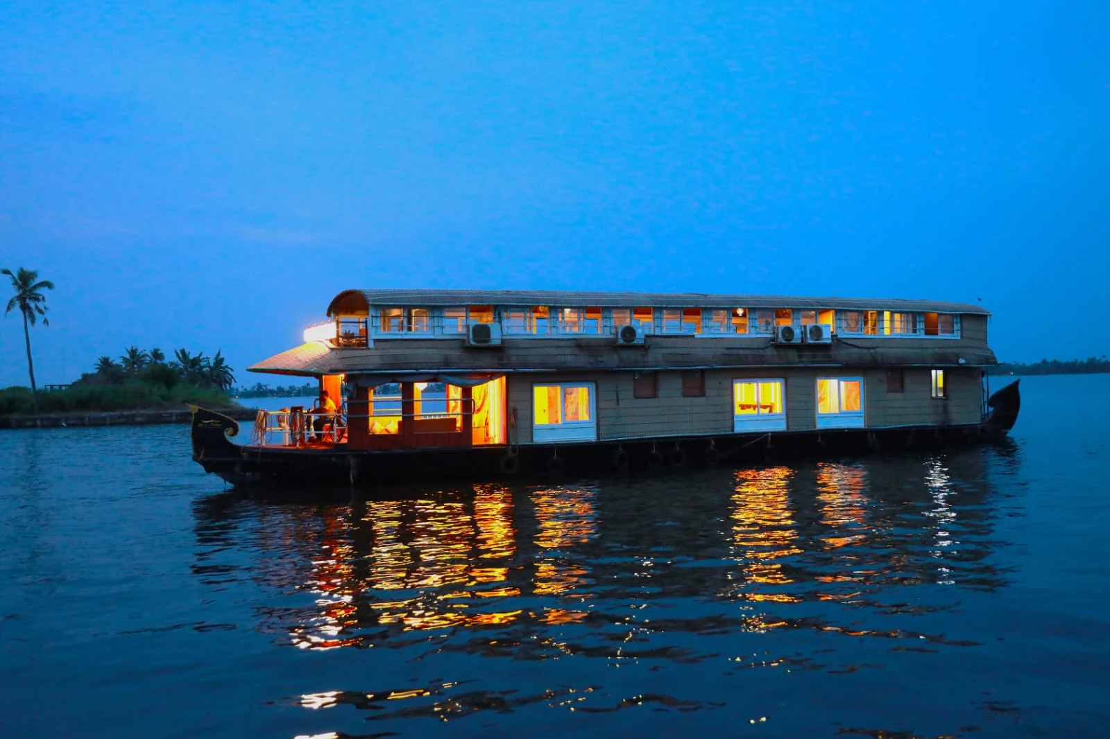A two-deck houseboat gliding across still blue water at dusk, its windows glowing amber and reflected in the lake