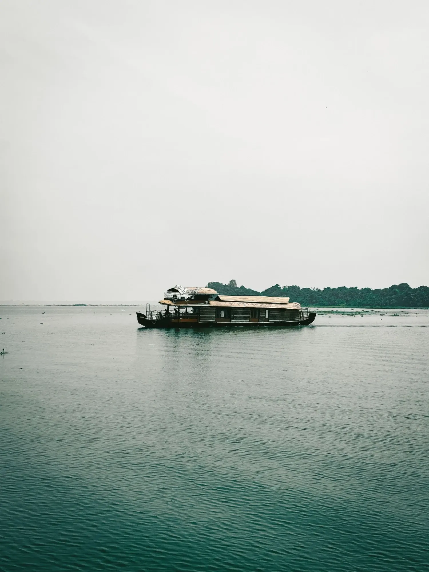 A houseboat cruising across a calm, glassy lake under a soft grey sky on the Alleppey backwaters