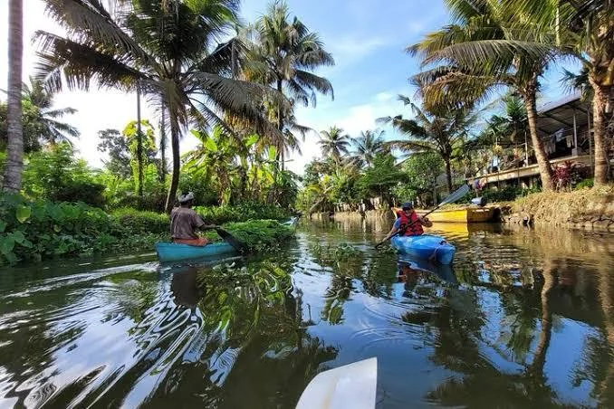 Two kayakers paddling a narrow, palm-lined Alleppey canal under a blue sky, with a village house on the bank