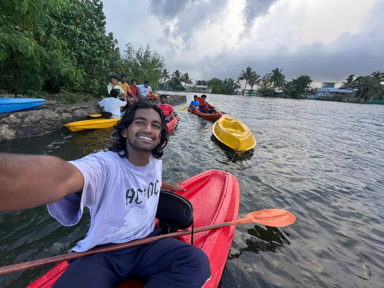 A smiling kayaker in a red kayak with a group of friends and more kayaks gathered at the lake's edge under a stormy evening sky