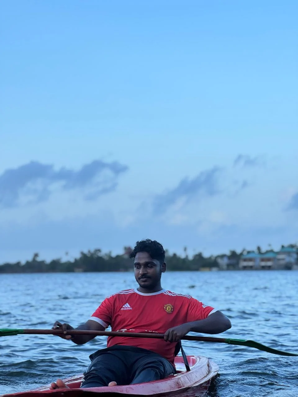 A kayaker in a red shirt paddling a red kayak on open backwater at dusk, with a calm blue sky behind
