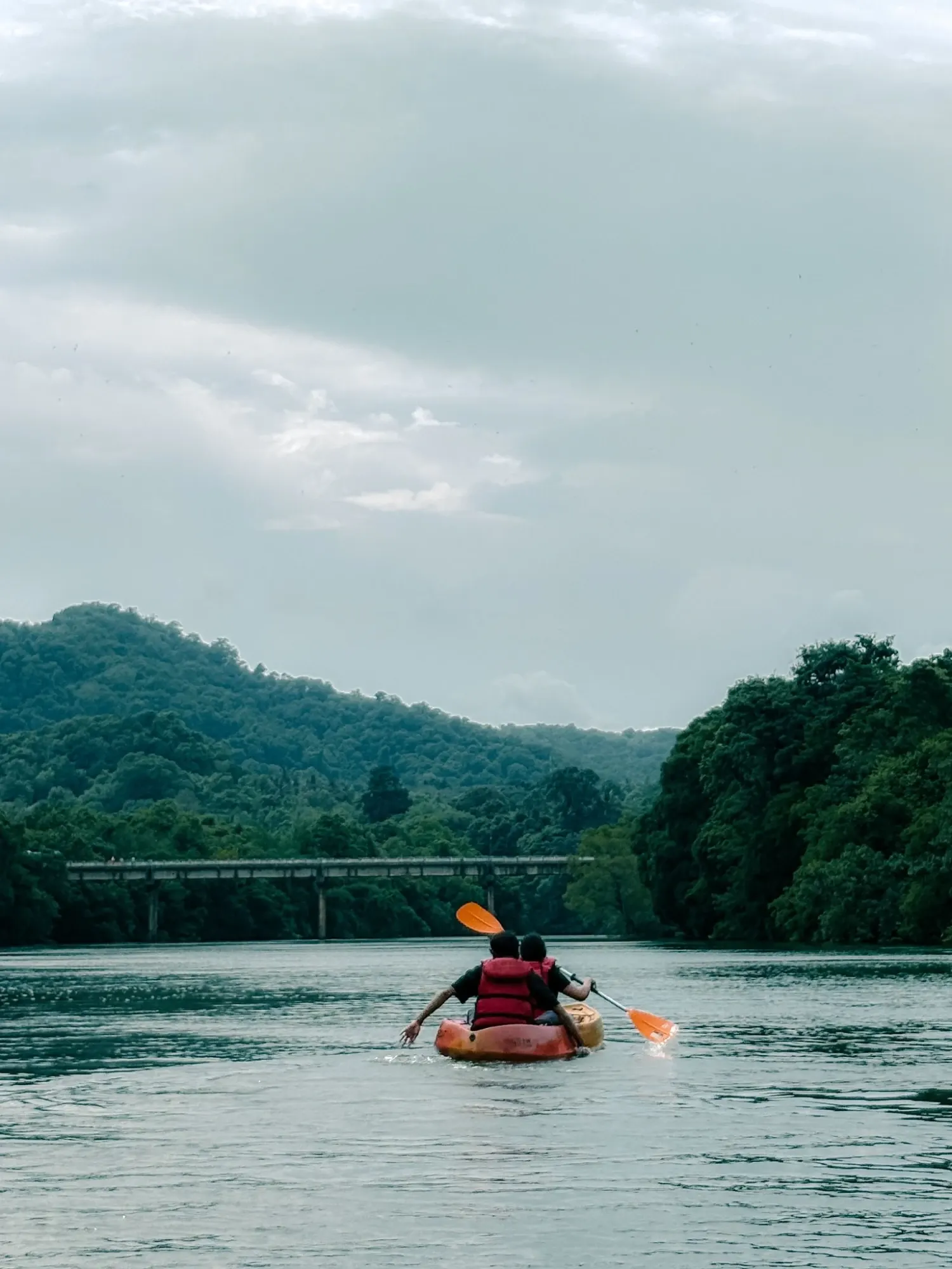 Two kayakers in red life jackets paddling an orange kayak on a calm river toward a bridge, with green forested hills under a cloudy sky