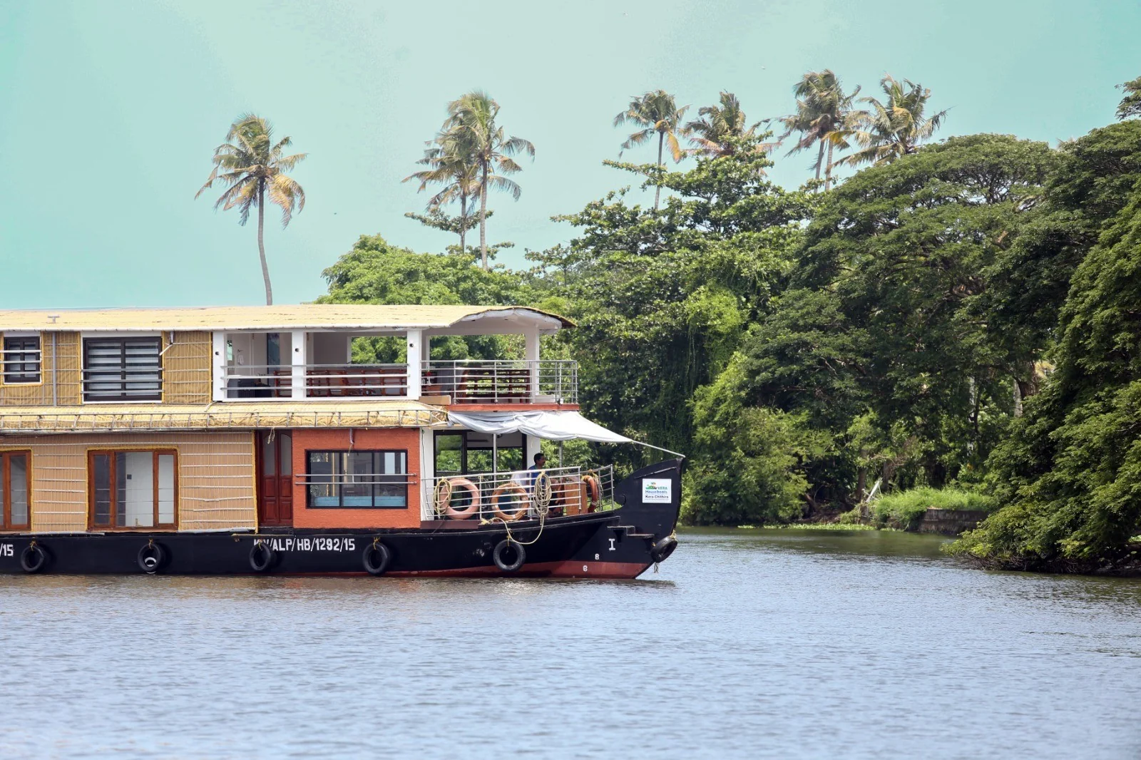 The four bedrooms Premium houseboat gliding along a tree-lined bank, with an open upper deck and thatched roofs