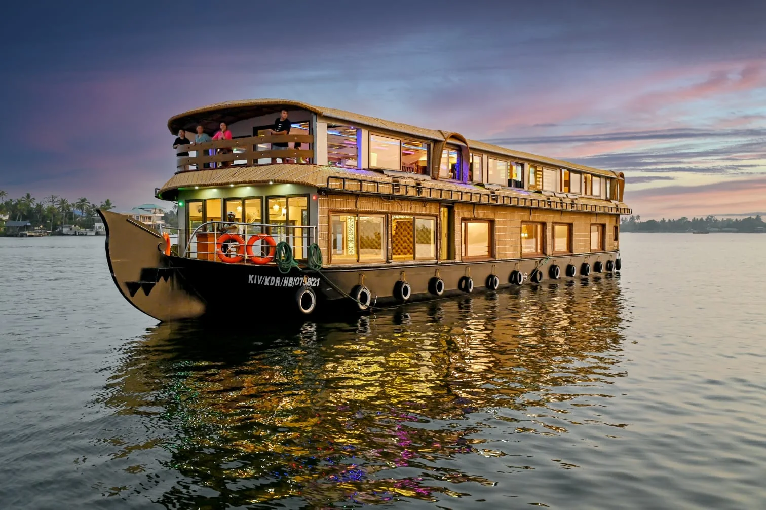 The seven bedrooms Premium houseboat on the water at sunset, with a long two-deck thatched body and a lit upper deck