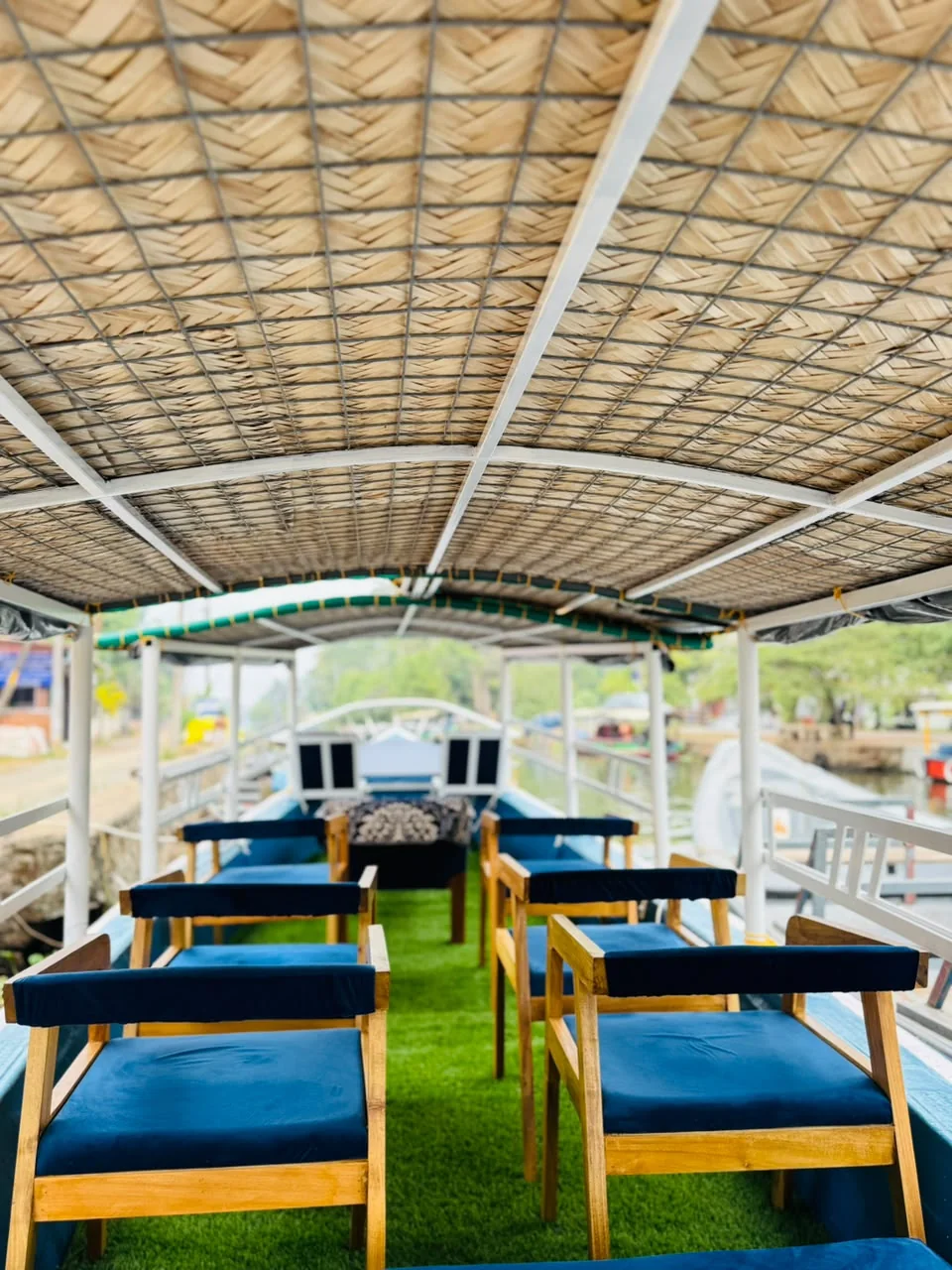 Rows of teak armchairs with navy cushions on green grass matting beneath a woven palm-leaf canopy