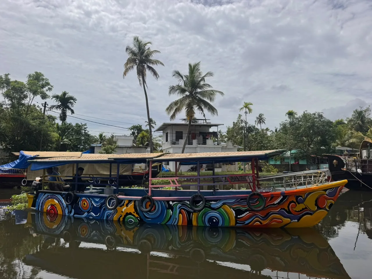 A hand-painted shikkara boat in swirling orange, blue and yellow, moored on a quiet backwater canal under tall coconut palms