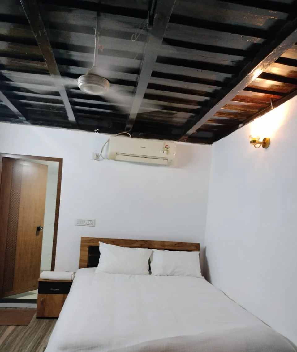 Bedroom with a white double bed, a wall lamp and a dark wooden-beam ceiling with a fan