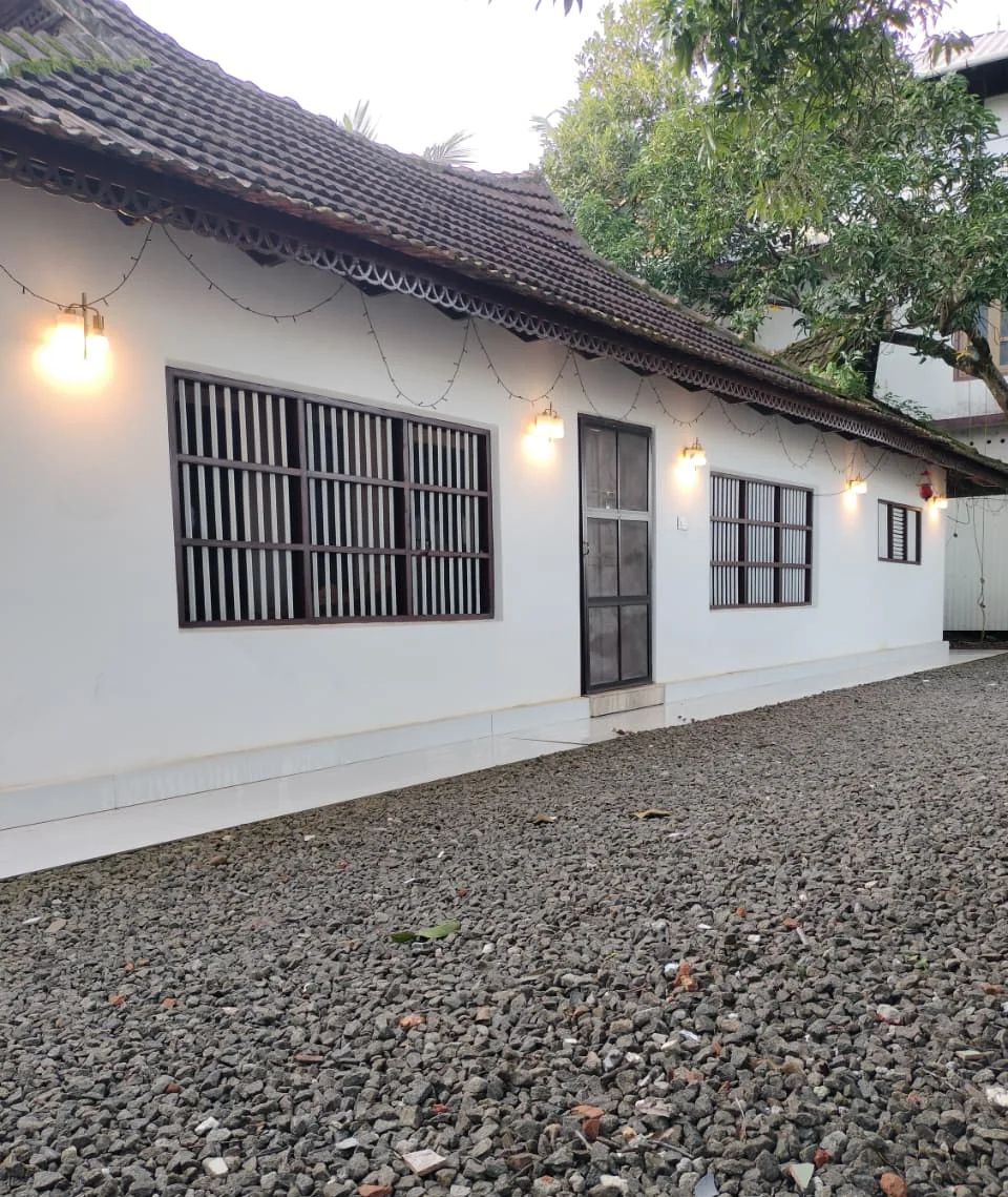 The front of a white Kerala heritage home with a tiled roof, carved eaves, timber-barred windows and warm wall lamps