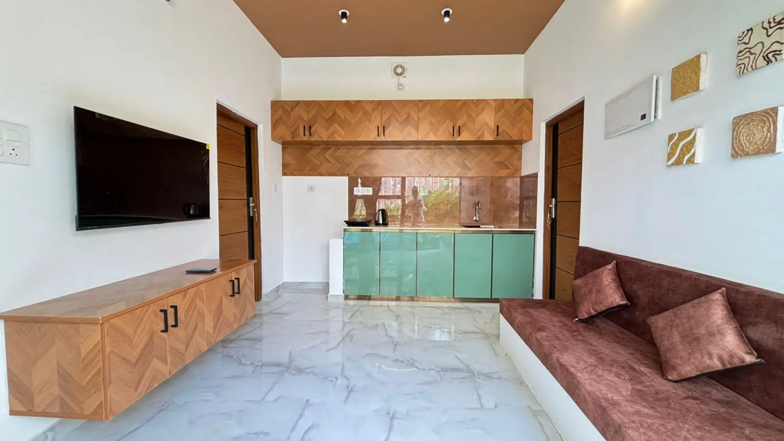 Open living area with a teak herringbone TV unit, a green kitchenette and a maroon built-in sofa on a marble floor